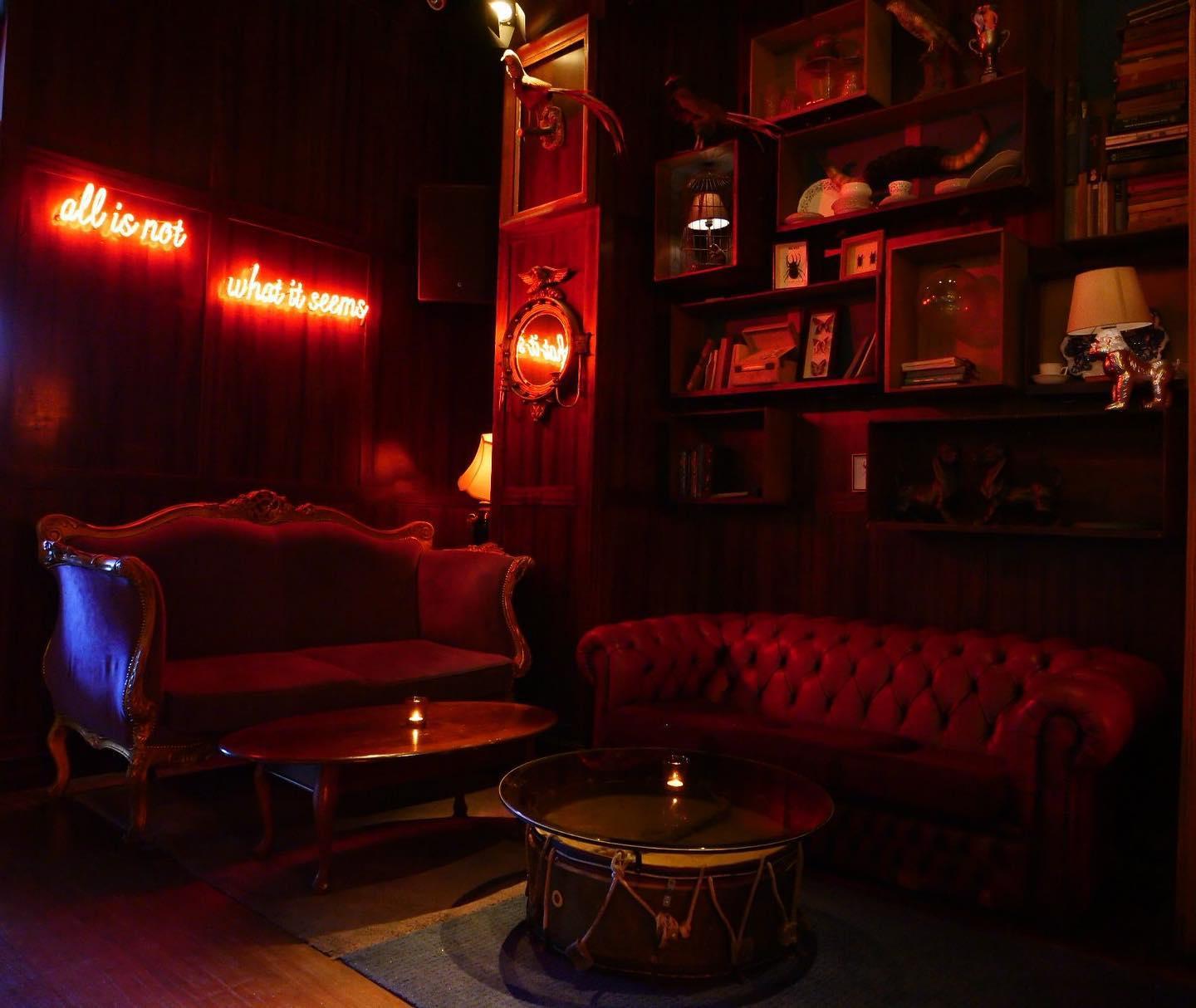 This is a cozy, dimly lit lounge area with vintage decor. On the left, there's a velvet sofa with ornate wooden details, and on the right, a deep red tufted leather couch. Between them, two round wooden tables hold small candles, with one table cleverly repurposed from a large drum. The walls are dark wood paneled and adorned with eclectic shelves displaying books, art, and curiosities, including bird and spider figurines. Warm, ambient light glows from table lamps and a neon sign on the wall that reads "all is not what it seems" in cursive orange-red script, adding a mysterious vibe to the room.