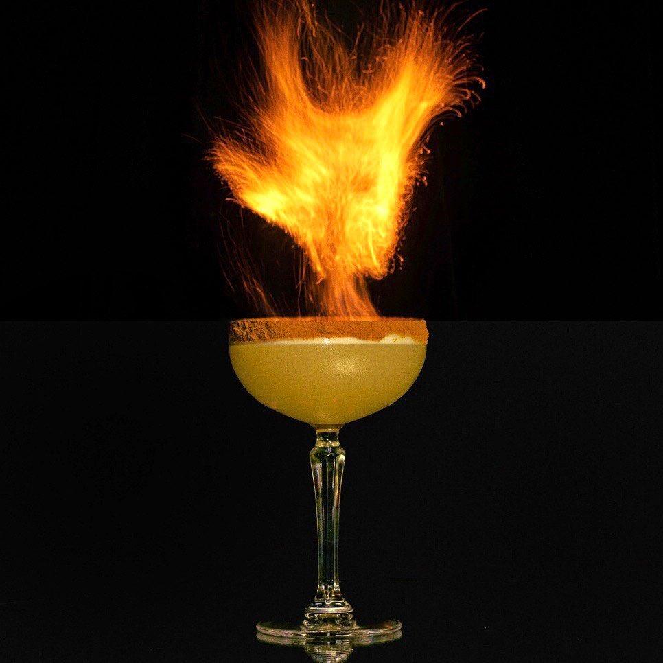 A cocktail in a coupe glass with a caramel or cinnamon-coated rim, set against a black background. A large, bright orange flame bursts dramatically from the top of the drink, creating a striking and dynamic visual effect.