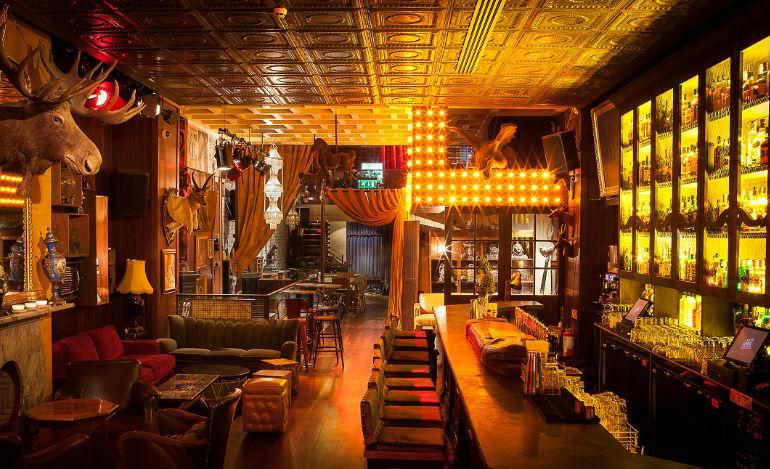 A cozy, warmly lit bar interior with vintage and rustic decor. The ceiling is patterned with tin tiles, and walls are adorned with mounted animal heads, including a large moose. There are plush sofas and armchairs arranged around low tables on the left side. The right side features a long bar with high stools, backlit shelves filled with bottles, and a large illuminated cross-shaped light fixture hanging above. The atmosphere is inviting with a blend of classic and eclectic elements.