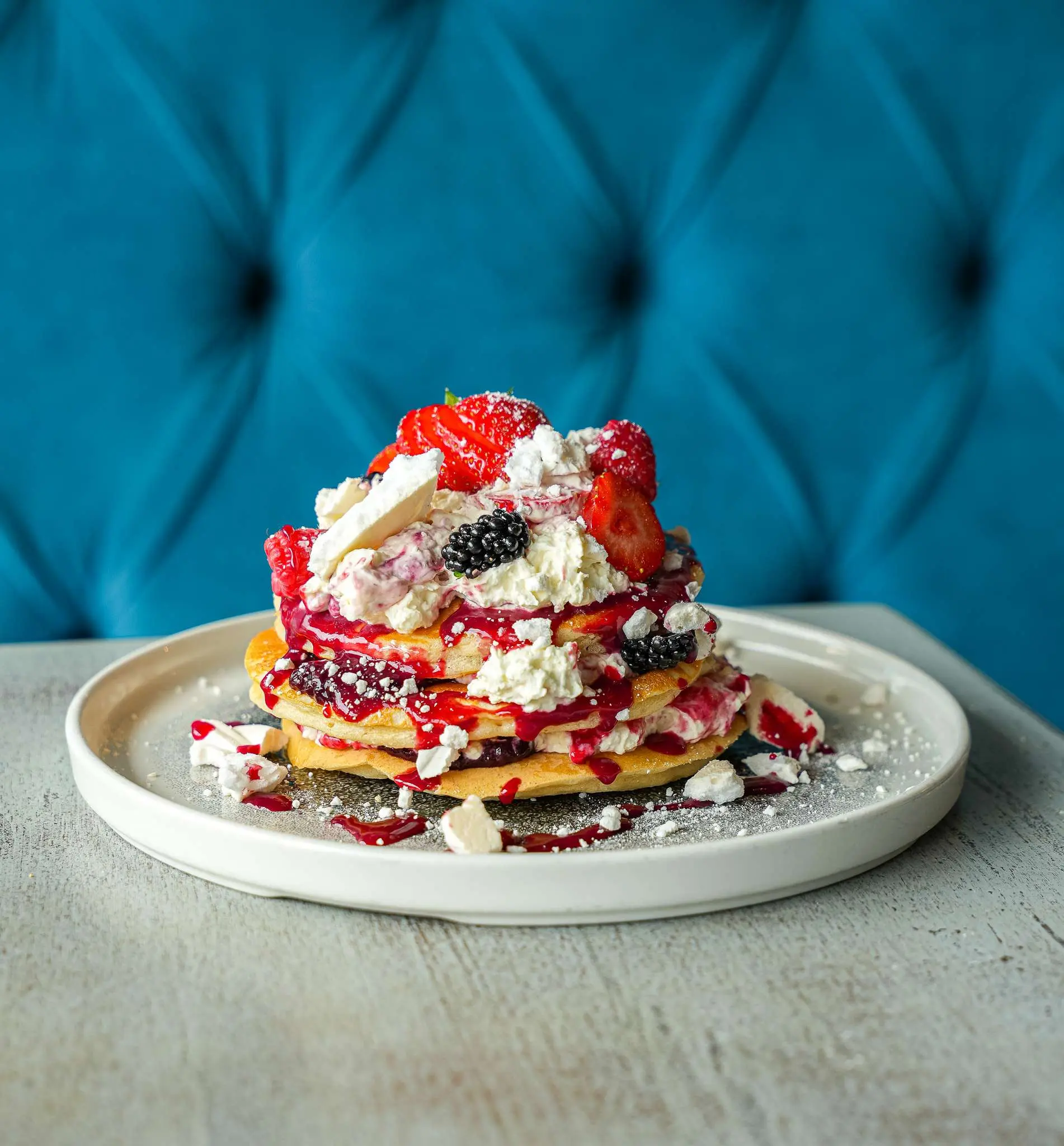 Stack of golden pancakes topped with whipped cream, strawberries, blackberries, berry sauce, and crushed meringue on a white plate.