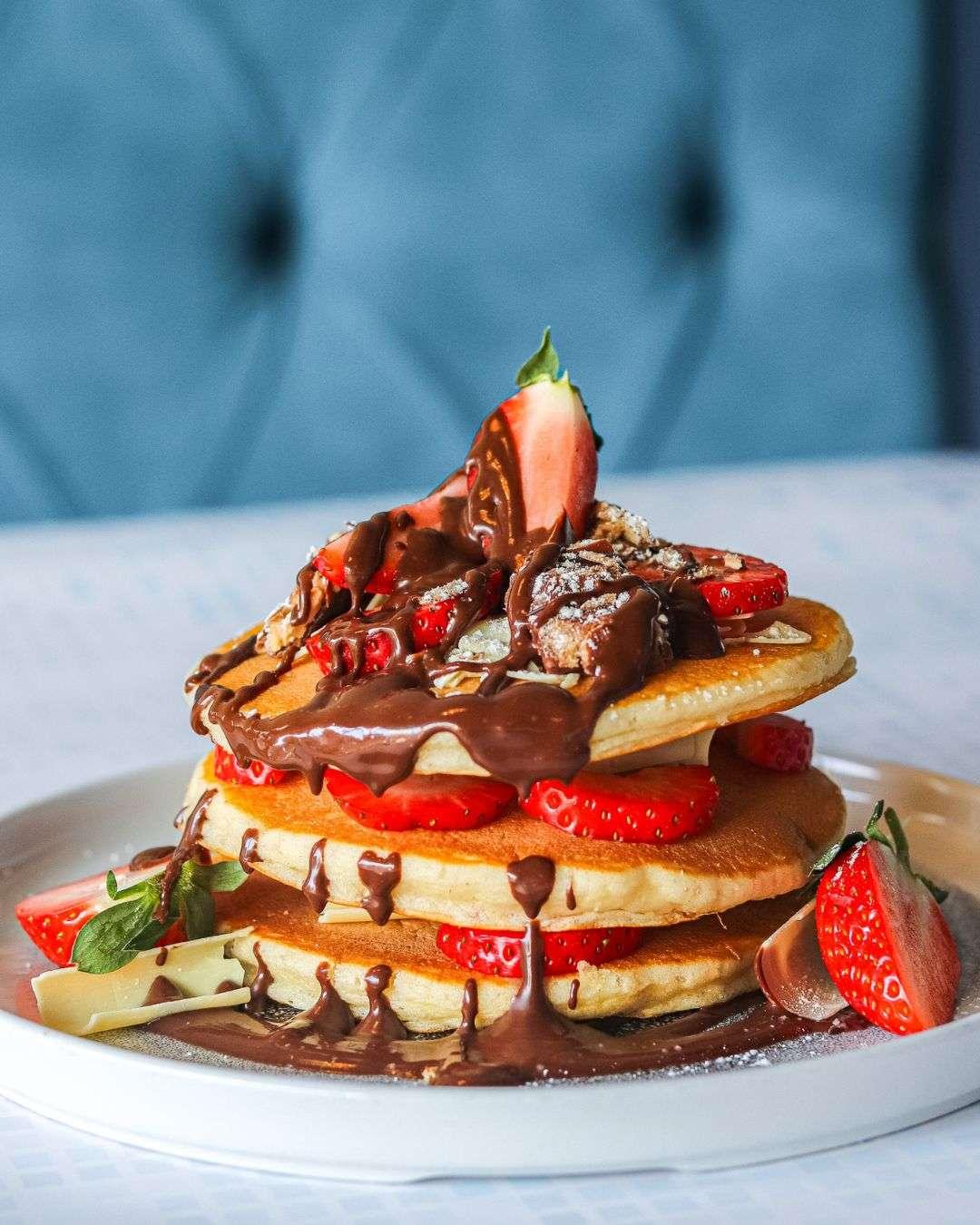 Stack of pancakes layered with fresh strawberries, drizzled with melted chocolate, topped with a halved strawberry and white chocolate pieces.