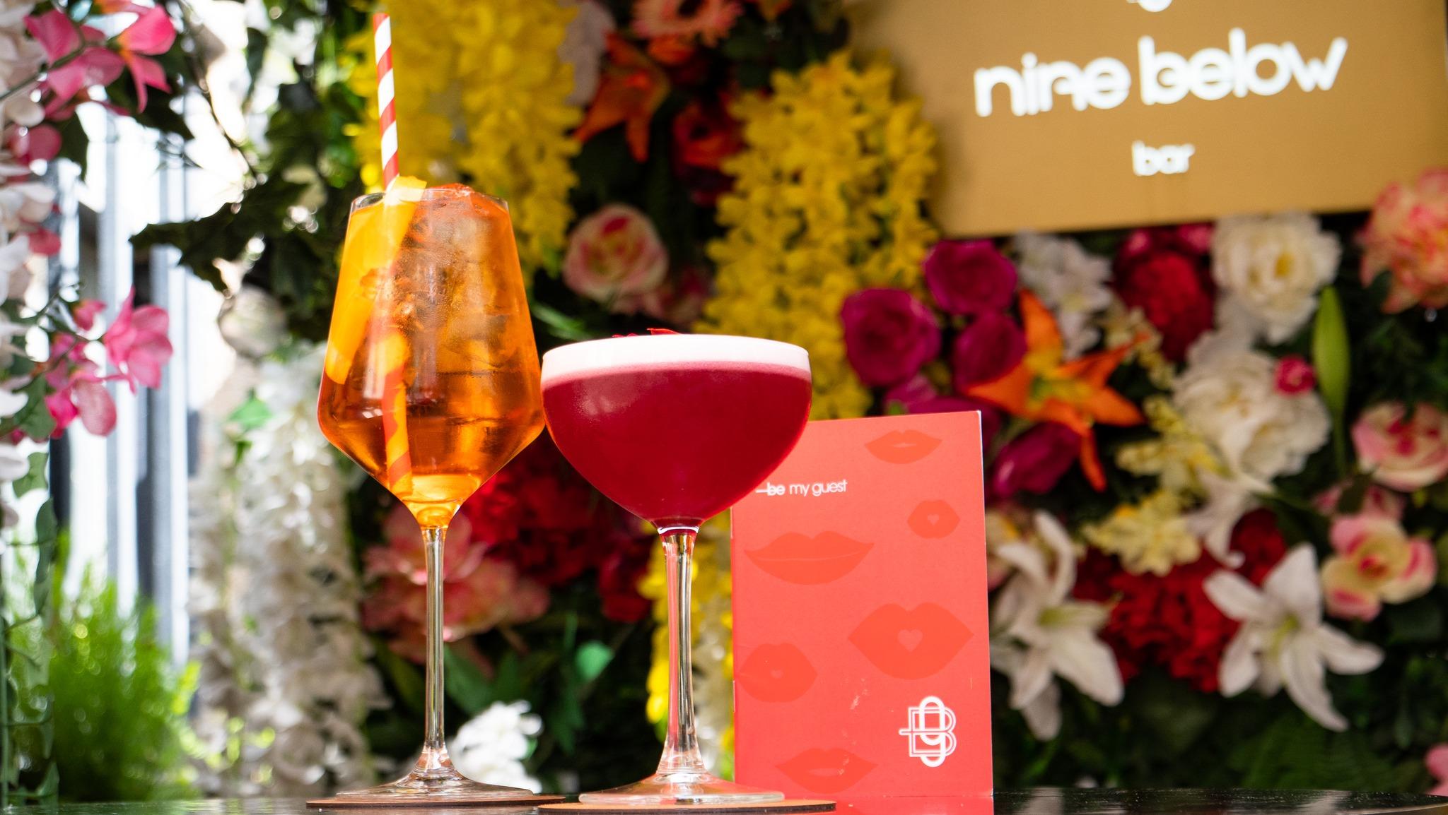 Two cocktails sit on a reflective surface in front of a colorful flower wall. The drink on the left is orange with ice and a red-and-white striped straw, garnished with a lemon slice. The drink on the right is a deep pink cocktail with a frothy white top. Between the drinks is a red card with lips and the text "—be my guest." In the background, a gold sign reads "nine below bar."