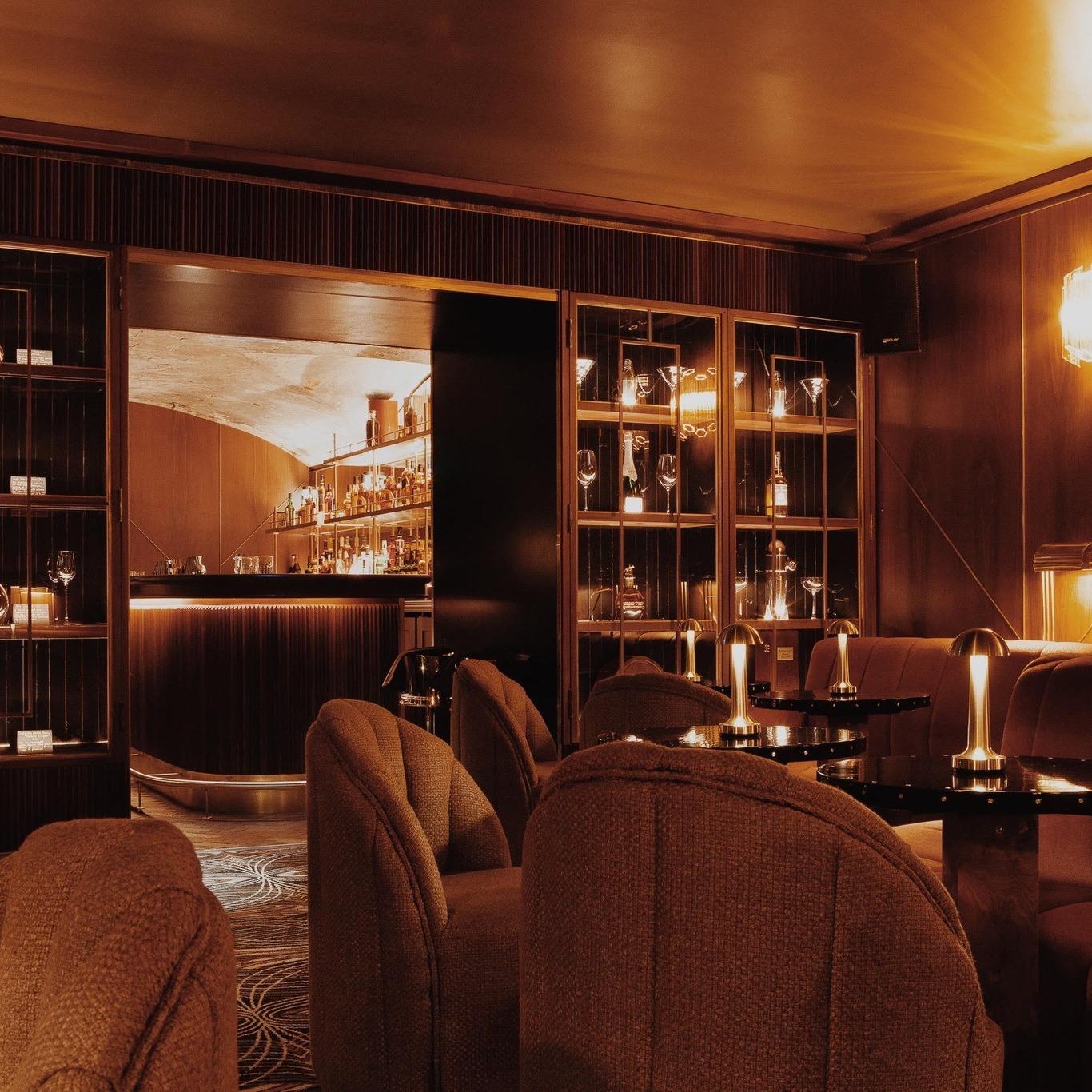 Cozy, dimly lit bar interior with plush upholstered chairs and black tables topped with warm golden lamps. A wooden and glass display cabinet holds glassware, while an open doorway reveals a well-stocked bar with bottles illuminated by soft lighting. The ambiance is warm with rich brown and golden tones throughout.