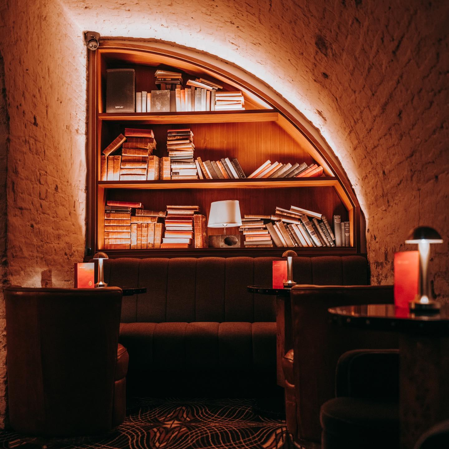 Cozy, dimly lit lounge area with a curved wooden bookshelf filled with books. The bookshelf is set into a brick archway with warm, ambient lighting outlining its shape. Below the bookshelf is a cushioned bench with two small round tables and leather armchairs on either side. Each table has a small lamp and a red menu or card.