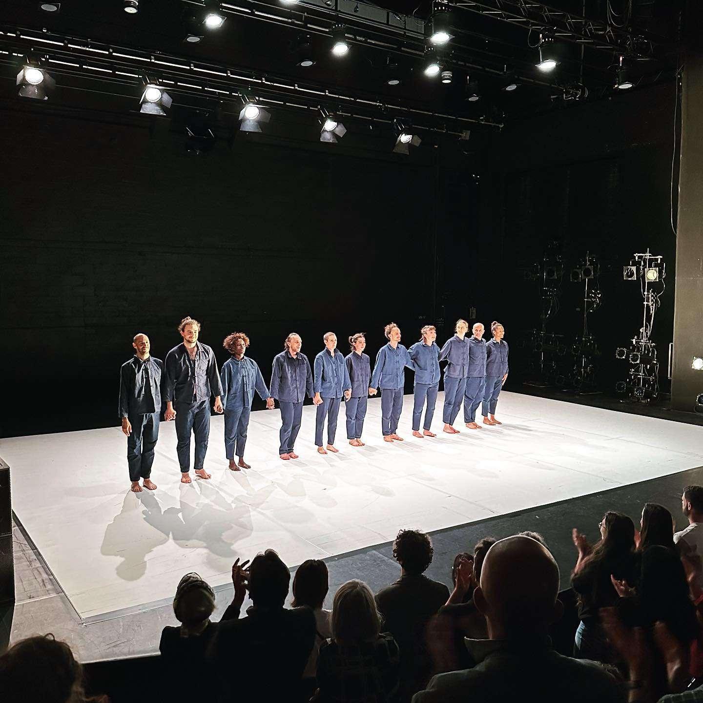 Ten performers stand barefoot on a brightly lit white stage, holding hands and facing the audience. They wear matching blue outfits and stand in a straight line. The audience, partially visible in silhouette, applauds them. The background is dark with stage lighting fixtures visible overhead and to the right.