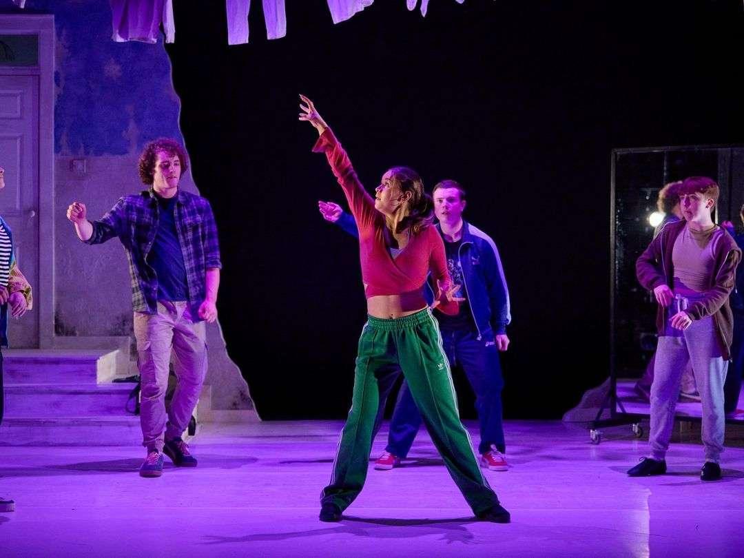 A group of young actors perform on stage under purple lighting. A woman in green track pants and a red cropped top stands center, reaching upward with her right arm. Around her, four men dressed casually in jackets, shirts, and pants move or gesture. The background is dark with a doorway and some hanging clothes visible on the left.