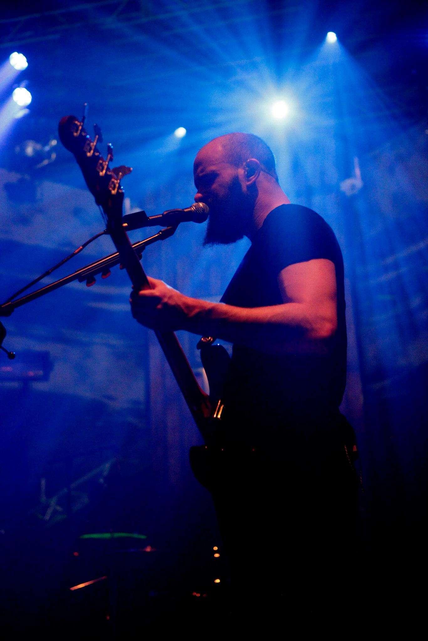 A bearded male musician wearing a black shirt playing an electric guitar and singing into a microphone on a dimly lit stage with blue and purple spotlights shining in the background.