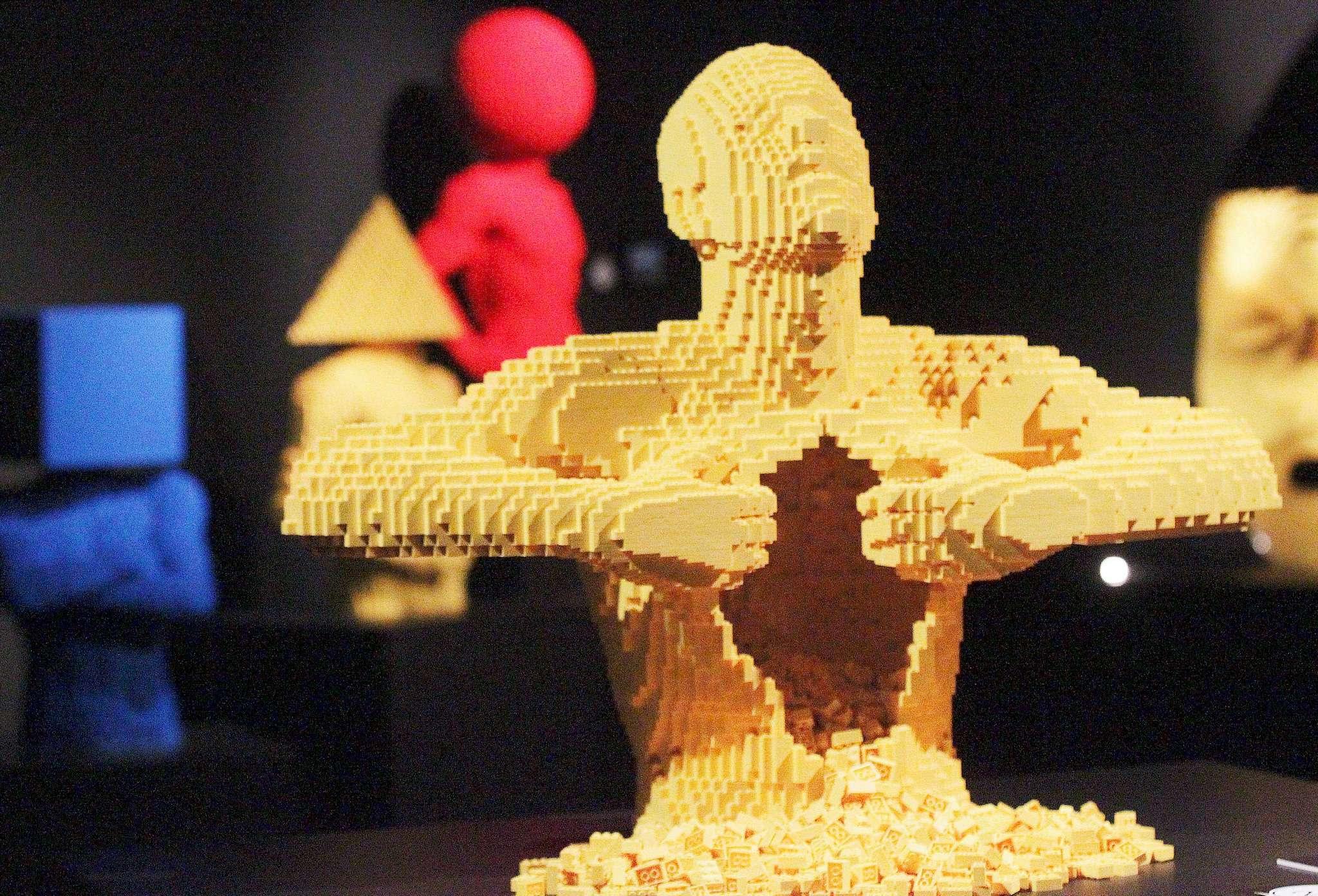 A yellow Lego sculpture of a human figure tearing open its chest, with Lego bricks spilling out. In the blurred background, there are other colorful Lego sculptures including a red figure, a blue figure, and a figure with a yellow conical hat.