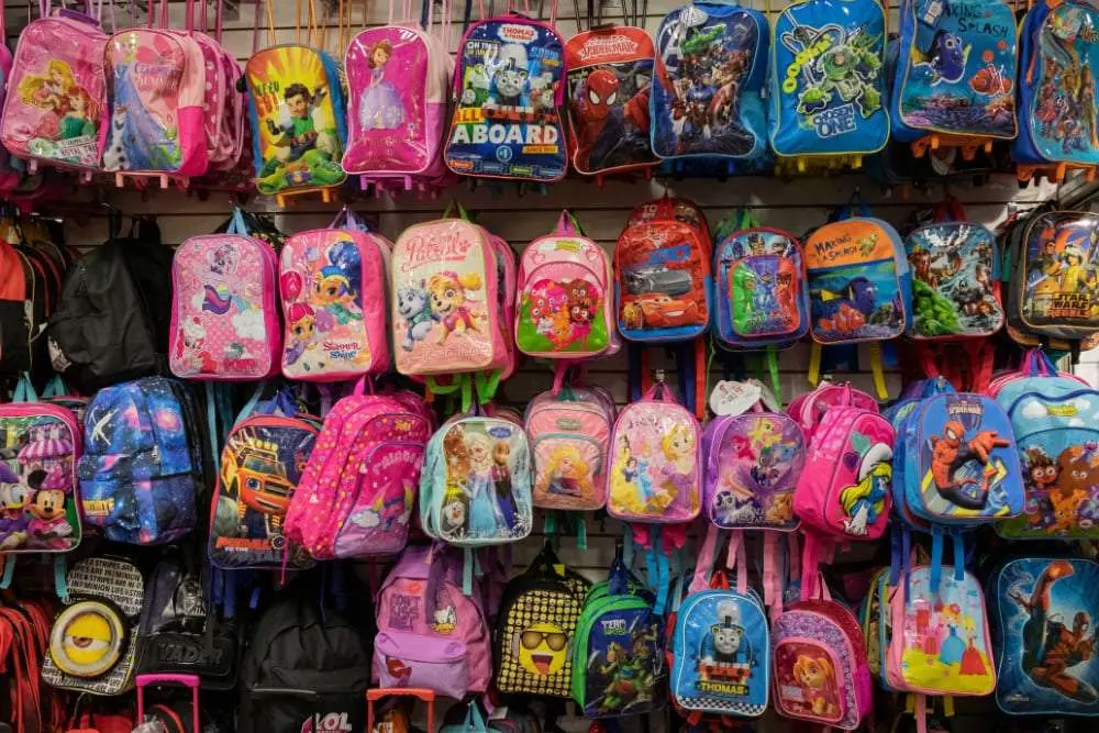 A store display of children’s backpacks in bright colors, decorated with popular animated and cartoon characters including Disney princesses, superheroes, and characters from shows like Paw Patrol, Frozen, and Thomas the Tank Engine.