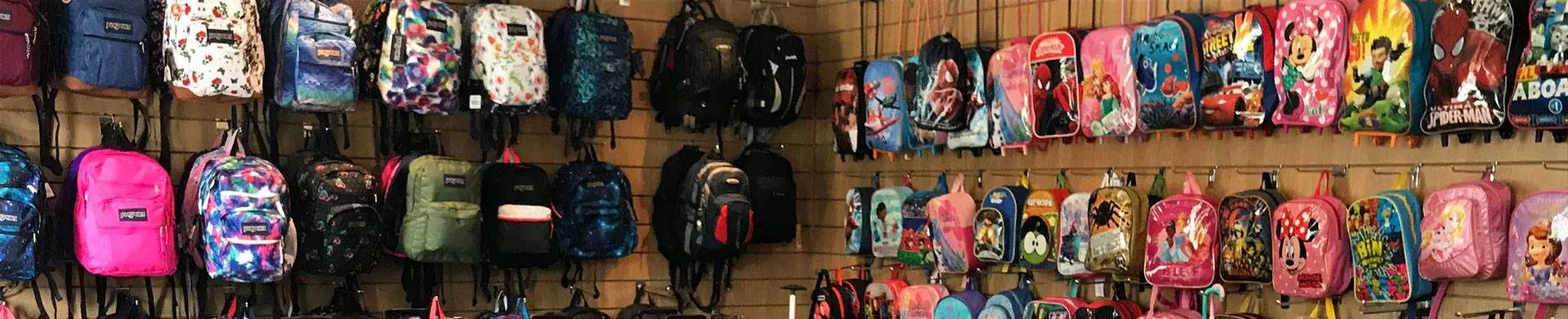 A retail display of various backpacks arranged on a wall, featuring solid-colored, patterned, and character-themed designs including superheroes, Disney characters, and animated figures.