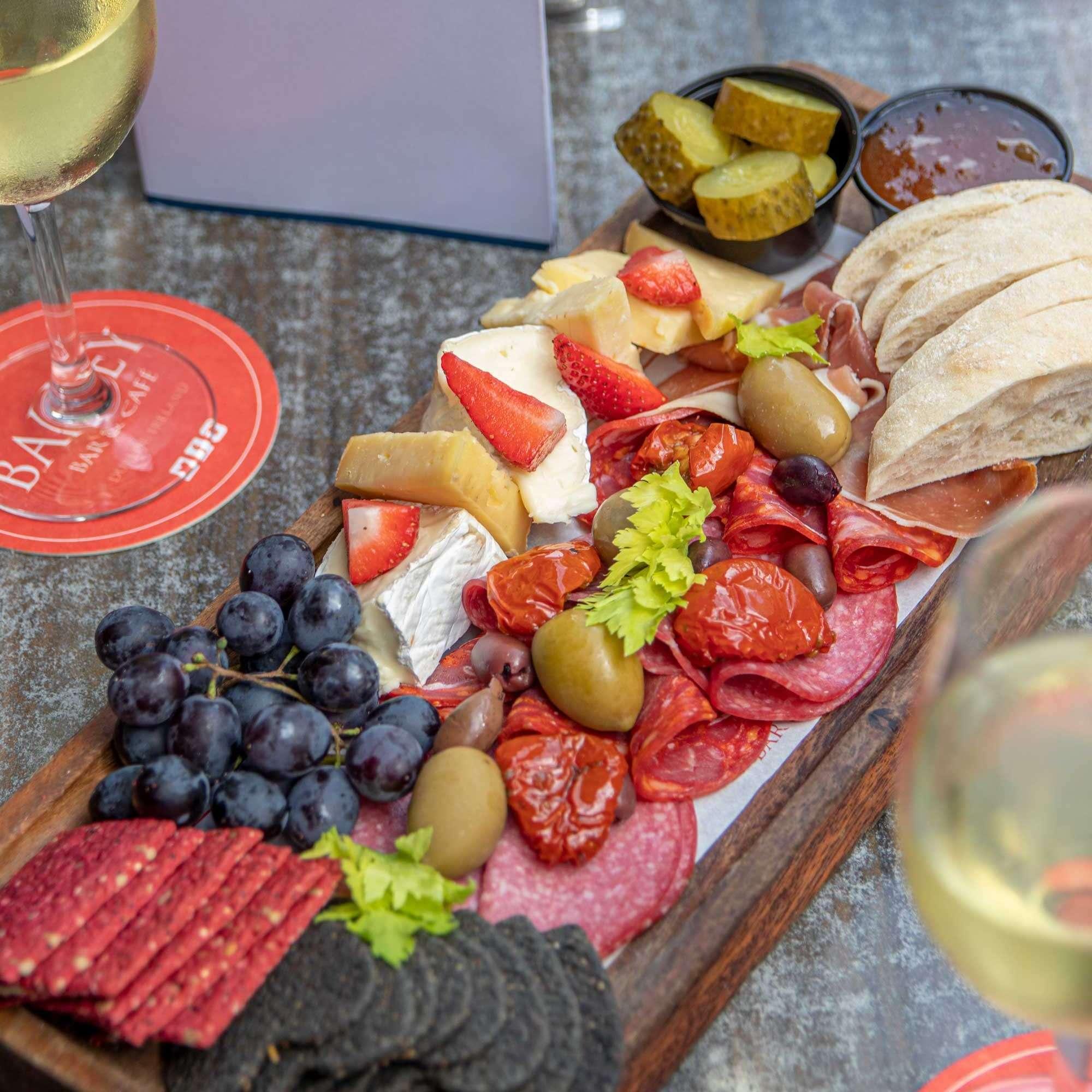 This image showcases a beautifully arranged charcuterie board served on a rustic wooden tray. The board includes an assortment of cured meats, cheeses topped with strawberries, pickles, black and green olives, sun-dried tomatoes, grapes, and artisan crackers. On the side, there are slices of crusty bread and small containers of jam and pickles. Two glasses of white wine and red Bailey Bar & Café coasters are also visible, suggesting the setting is a relaxed outdoor dining area, perfect for sharing and socializing.