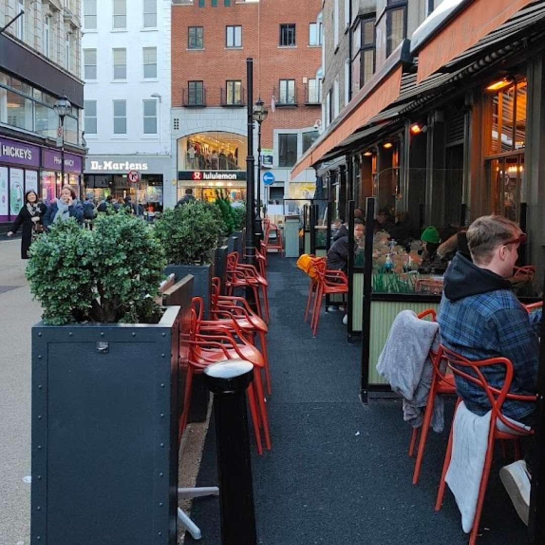 This image shows a lively pedestrian street with outdoor seating along a café or restaurant. Bright red chairs line the sidewalk next to planters and a row of tables, where people are dining and chatting. In the background, well-known storefronts can be seen, adding a vibrant urban shopping vibe.