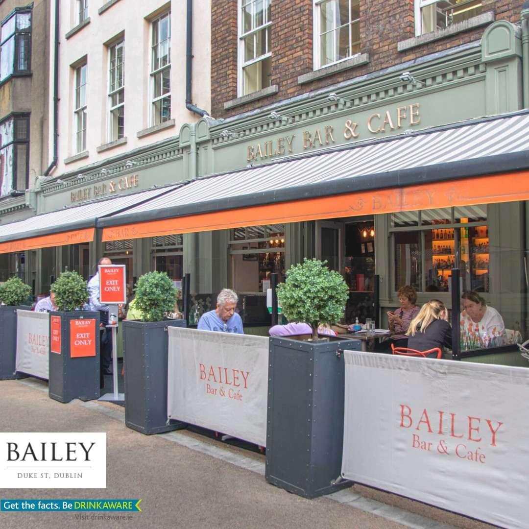This image shows Bailey Bar & Cafe on Duke Street, Dublin. The exterior is green with gold lettering, and there’s outdoor seating under orange-striped awnings. People are dining outside, separated from the sidewalk by planters with shrubs. The atmosphere looks inviting and lively.