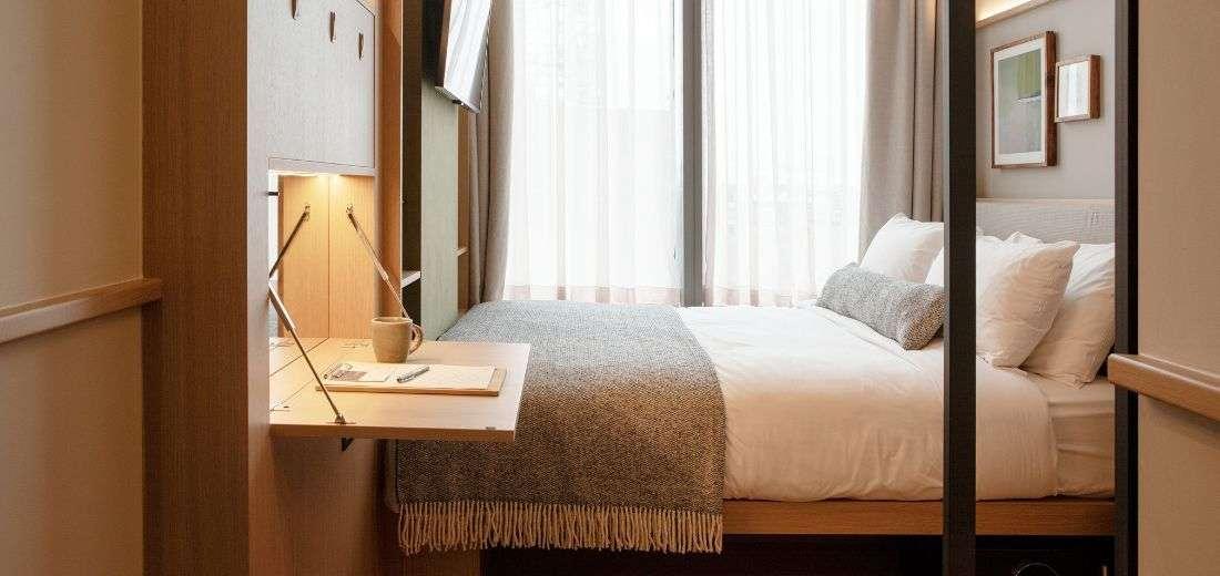 Compact hotel room with a neatly made double bed, a mounted TV, a small wall-mounted desk with a cup and notepad, and floor-to-ceiling sheer curtains letting in natural light.