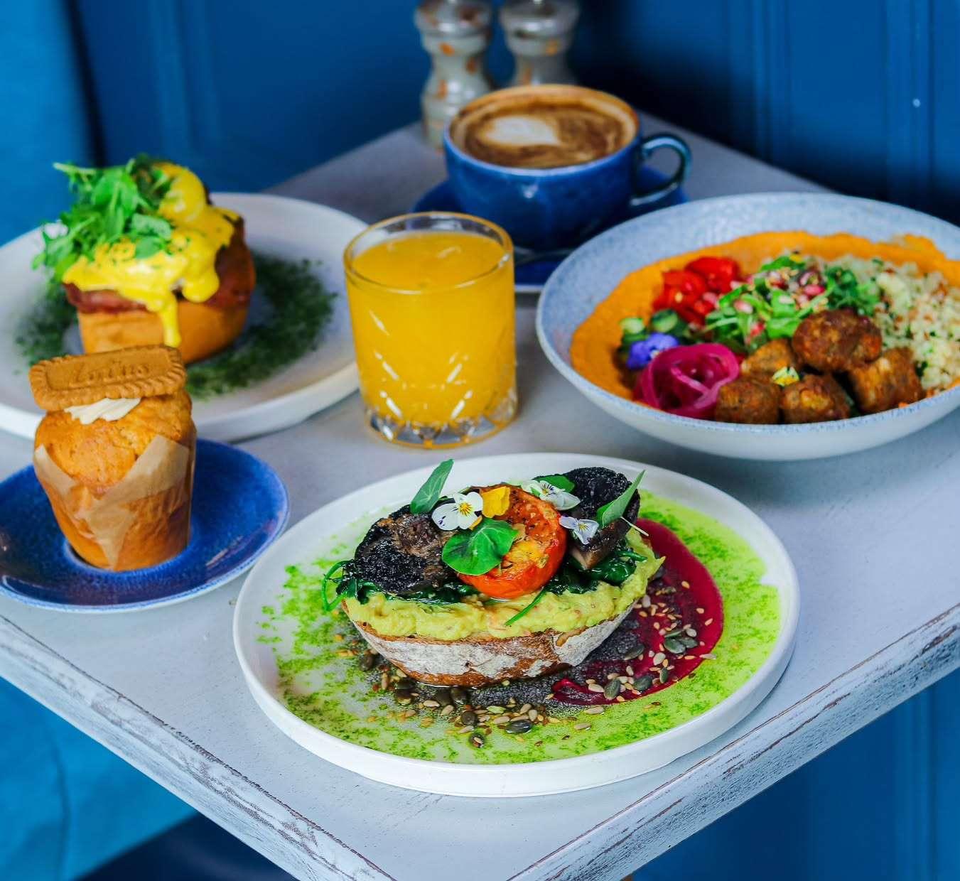 Assorted brunch dishes including avocado toast with vegetables and edible flowers, a poached egg muffin with hollandaise, a grain bowl with falafel, a muffin with a biscuit, orange juice, and a cappuccino on a white table.