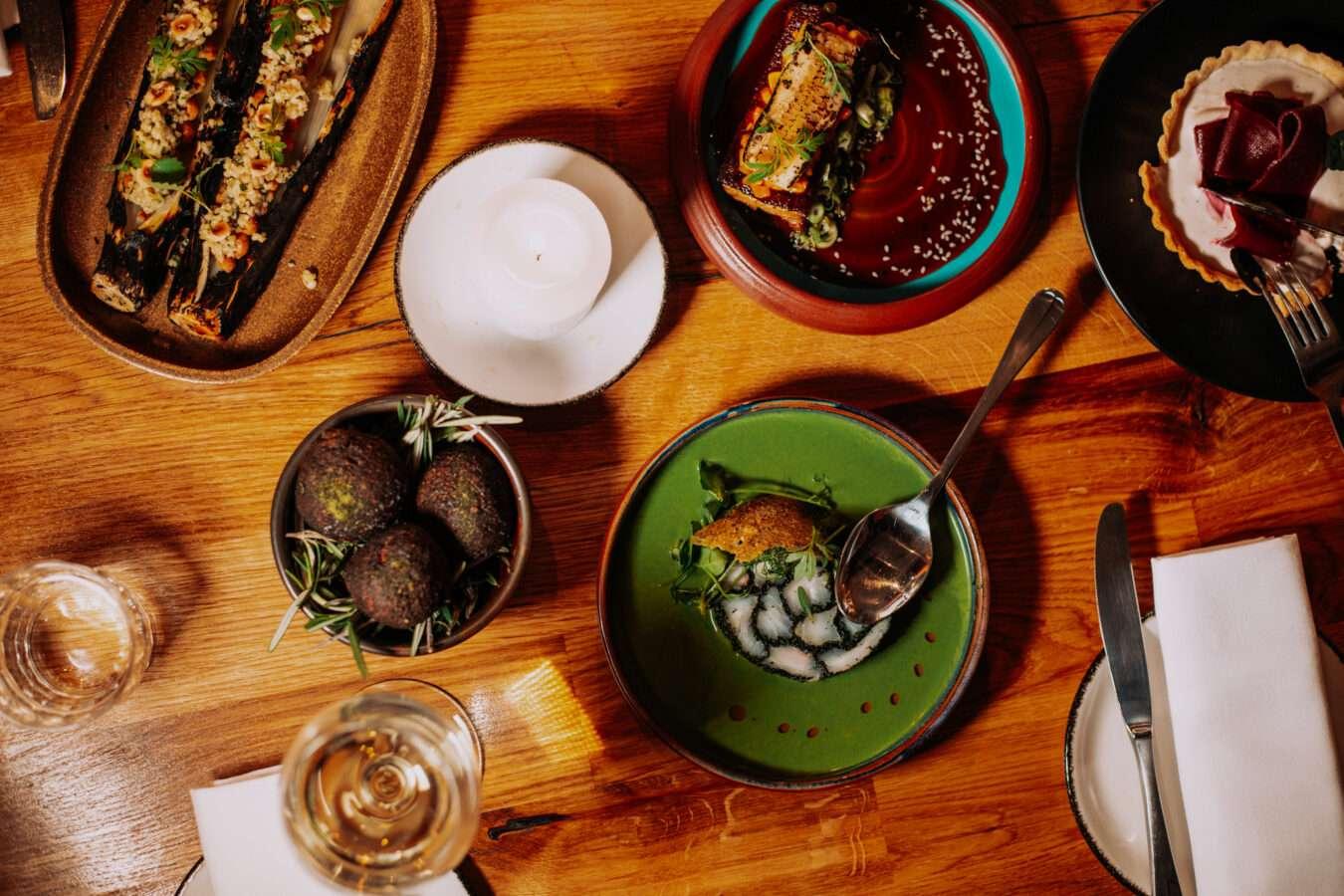 A top-down view of a wooden dining table with an assortment of colorful dishes including charred vegetables, green soup, meat or fish with sauce, truffle-like balls, and a dessert tart, with a lit candle in the center.